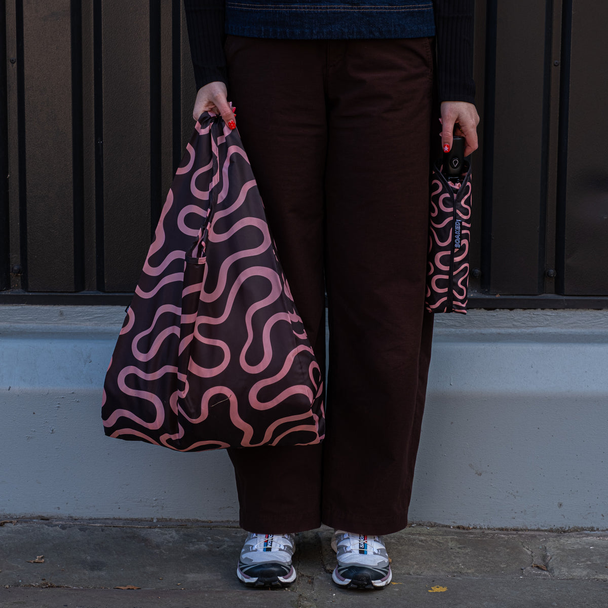 Person standing outside holding the SOAKED Doodle black and peach swirl tote bag open and the umbrella in its sleeve